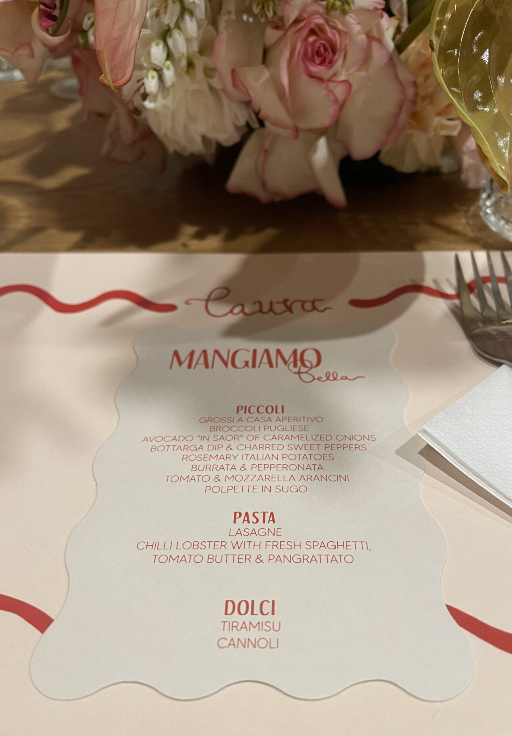 A personalised place setting for Laura, with a scalloped white menu with red writing. In the background are florals provided by Coral & Co., partners, sitting in the middle of the table.