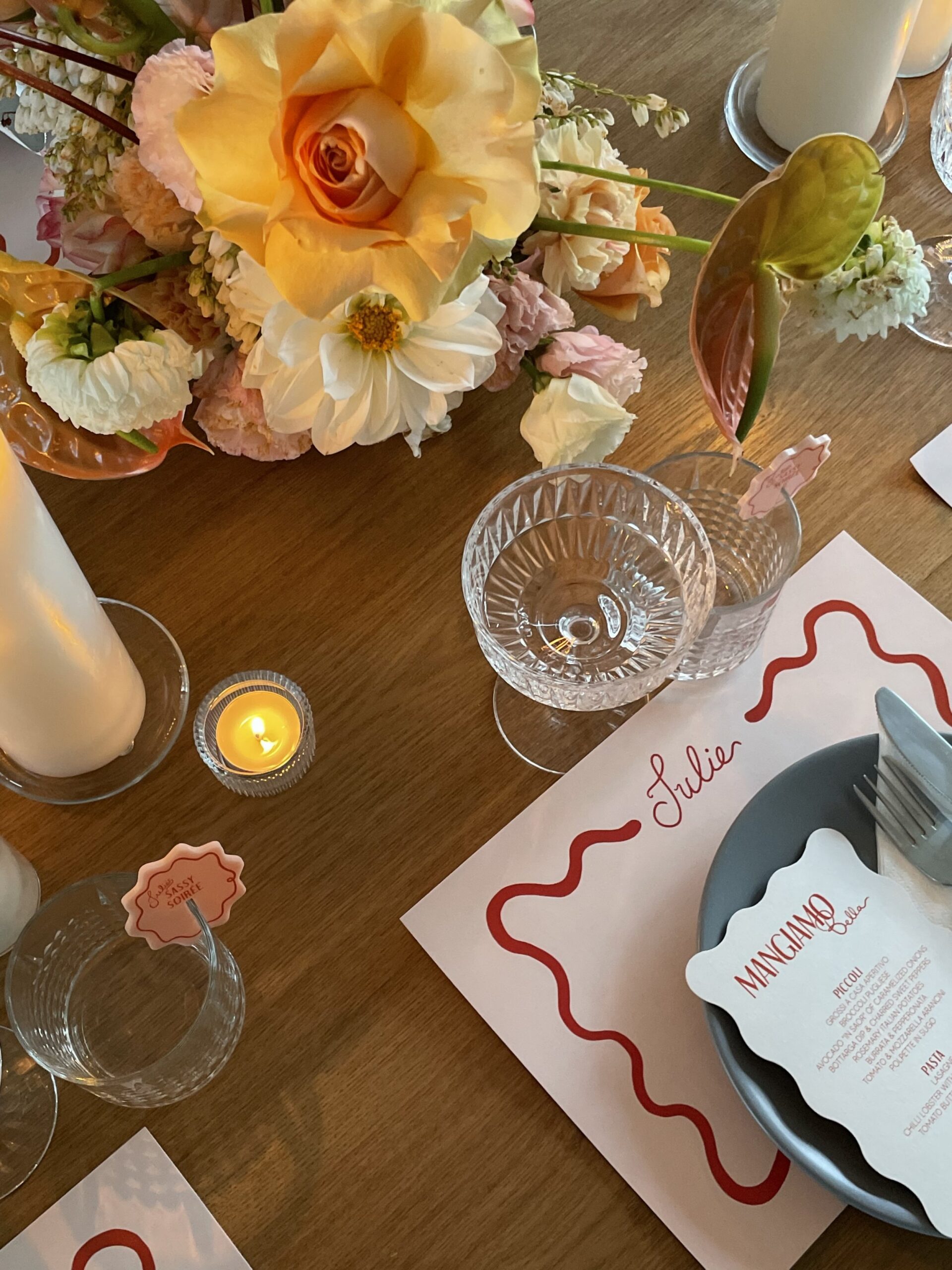 A table setting styled by Coral & Co., with scalloped white menu, with red writing sitting on a plate with cutlery, on a light pink rectangle place setting with the name "Julie" in red.  Wine glasses and water glasses with drink tags above the place setting and colourful  florals in a peach palette sit in the middle of the table. 