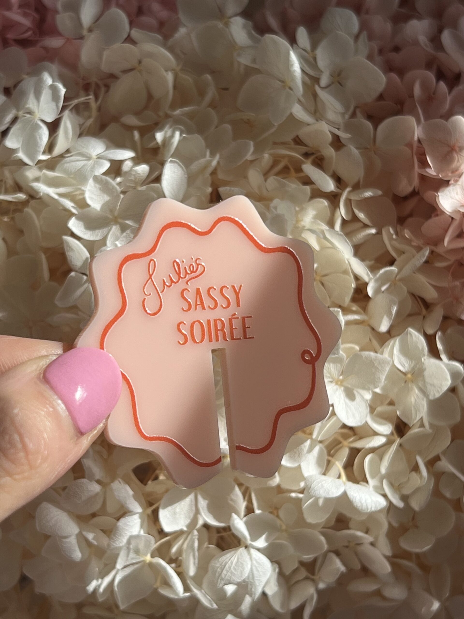 A ladies hand with bright pink nail varnish holds a personalised peachy pink drinks tag, with a wiggly outline and red wiggly border. In the middle of the tag reads "Julies Sassy Soiree". Designed and curated by Melbourne event stylist Coral & Co. 