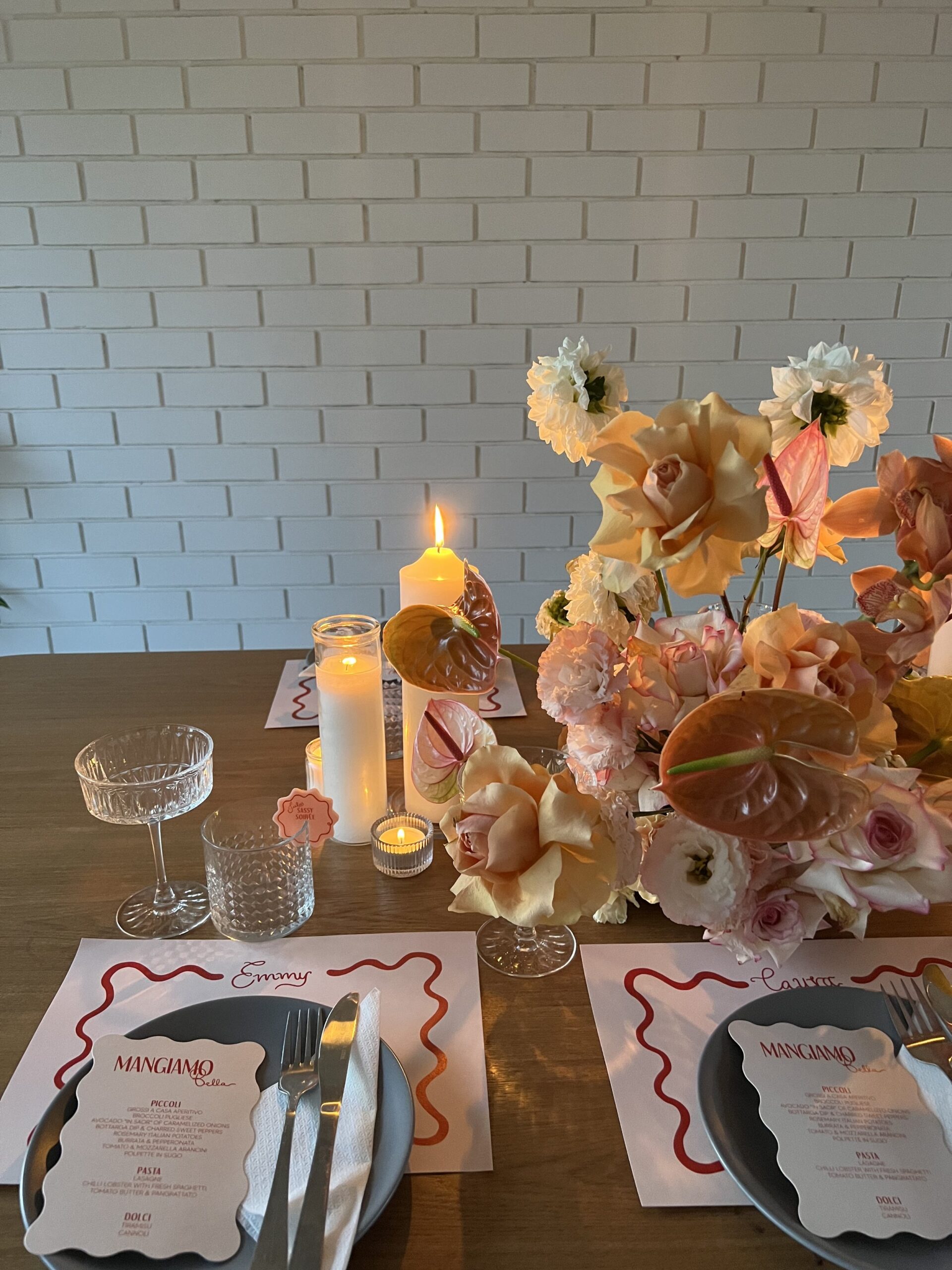 Two personalised table settings, with full, peach coloured florals, sitting next to a brightly burning tall candle and three smaller ones. Two more table settings are opposite, hidden by the large florals, and a white brick wall sits behind the table.