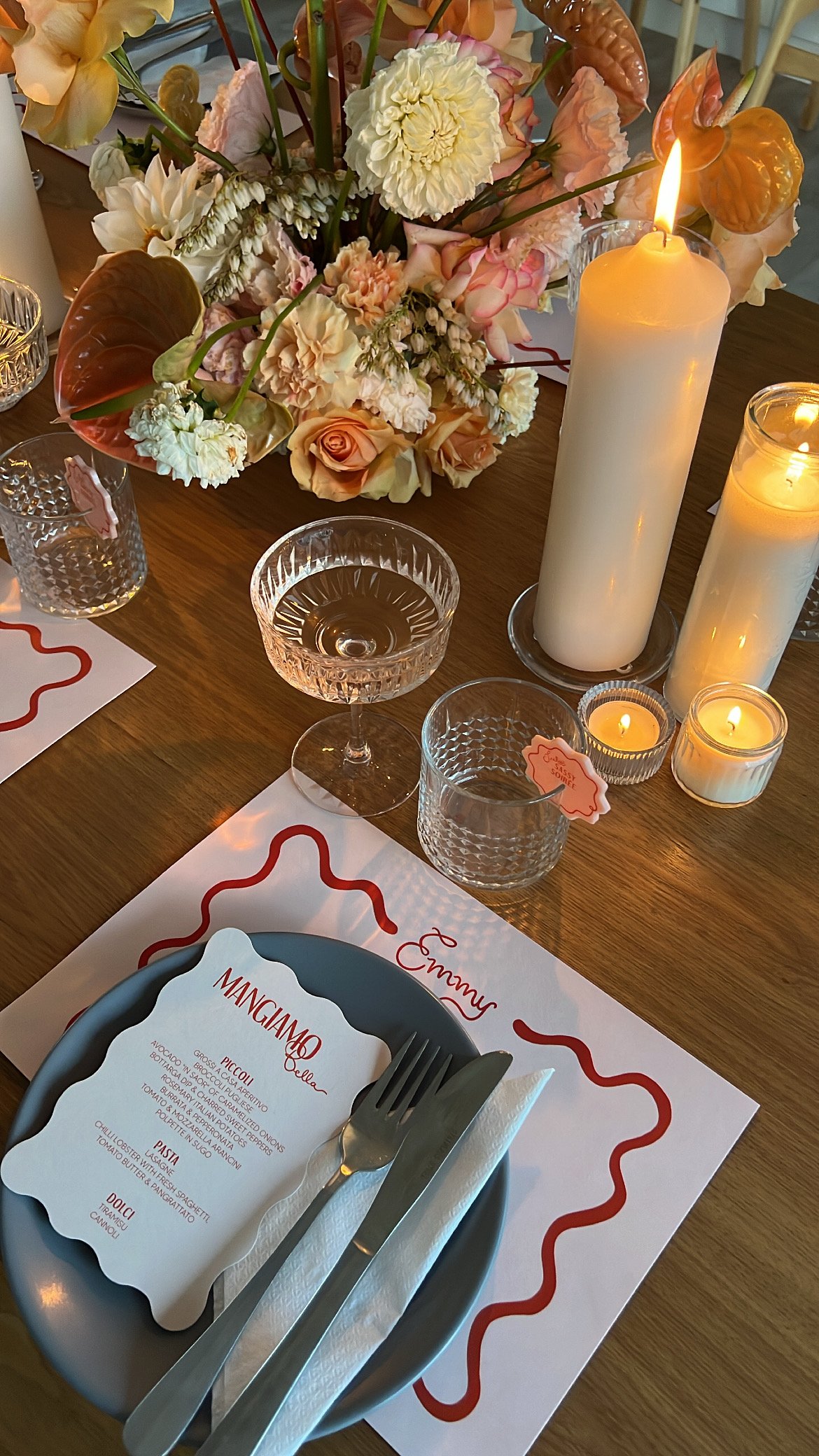 A personalised place setting for Emmy, with plate, menu and glasses. Four different sized white candles, in clear glass holders burn above the place setting. A full and vibrant floral accompanies the setting in the middle of the table. A Melbourne event by Coral & Co. 