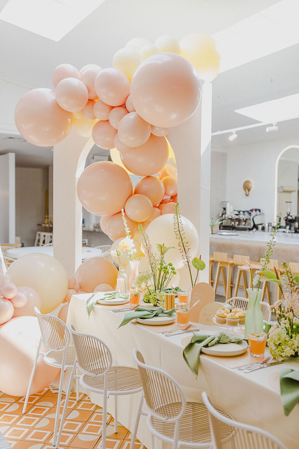 A bright table setting. Four chairs face a full table with avocado green tied napkins on plates, orange cocktails, green florals down the middle of the table and an arch next to the table. A flowing light peach and cream balloon garland stretches past the table.
Nothing says ‘celebration’ like a pretty pastel balloon arch! Blowout Balloons hit the brief with their perfectly colour matching, free flowing, breath-taking balloon arch that flowed effortlessly throughout the space.