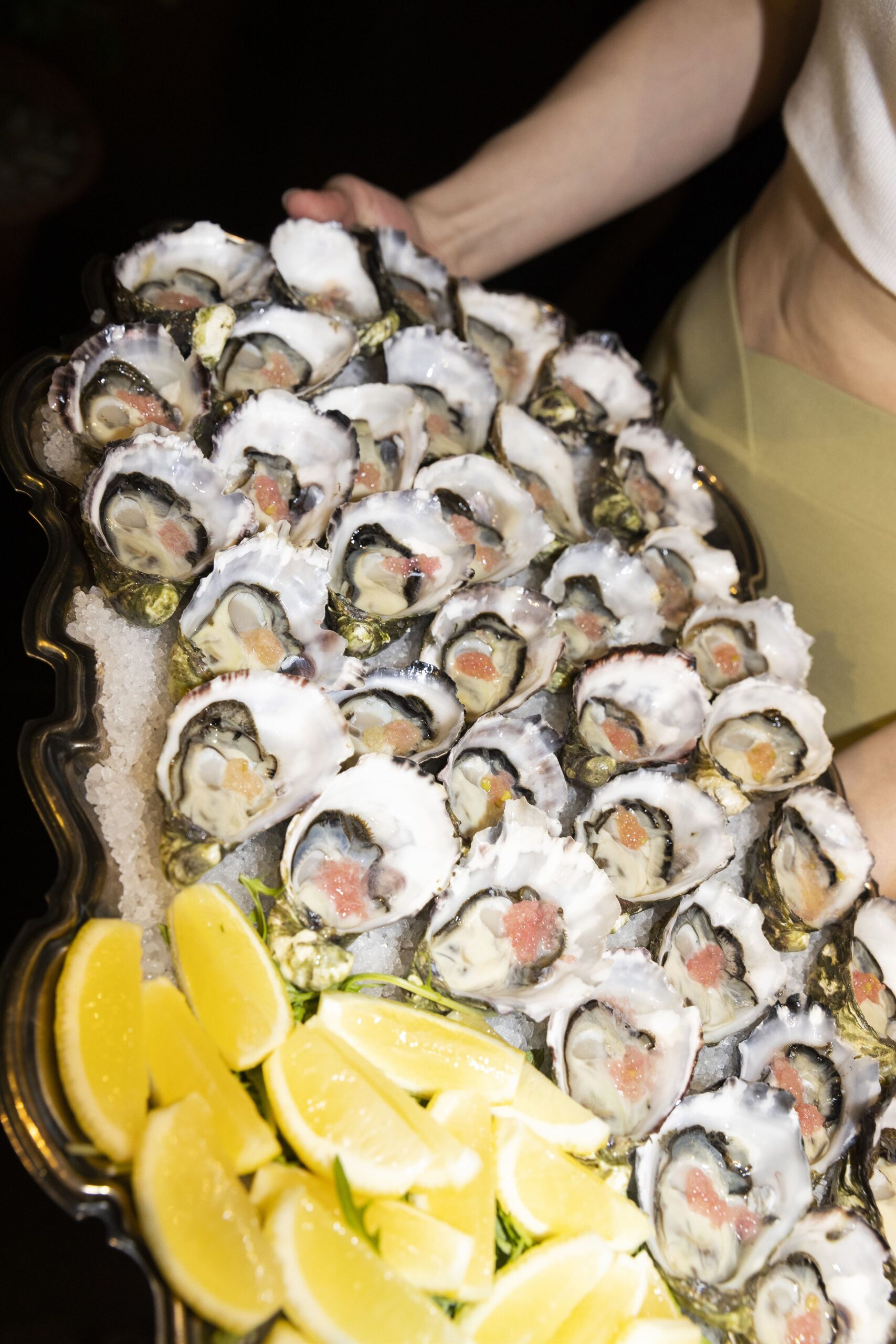 A platter of shucked oysters, sitting on a bed of salt, garnished and served with bright yellow wedges of lemon. 