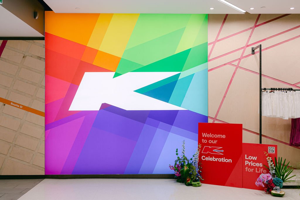 Bright Kmart sign, with large white K, surrounded by colours of the rainbow. Two welcome sign sit to the right of the large sign, with bright florals either side of the signs. Designed by Coral & Co., in Melbourne. 
