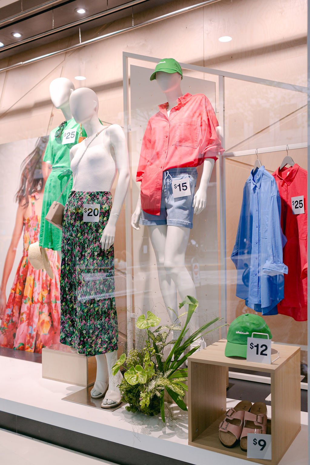 Window display at Kmart showcasing their new summer season range. Green florals sit between dressed mannequins. 