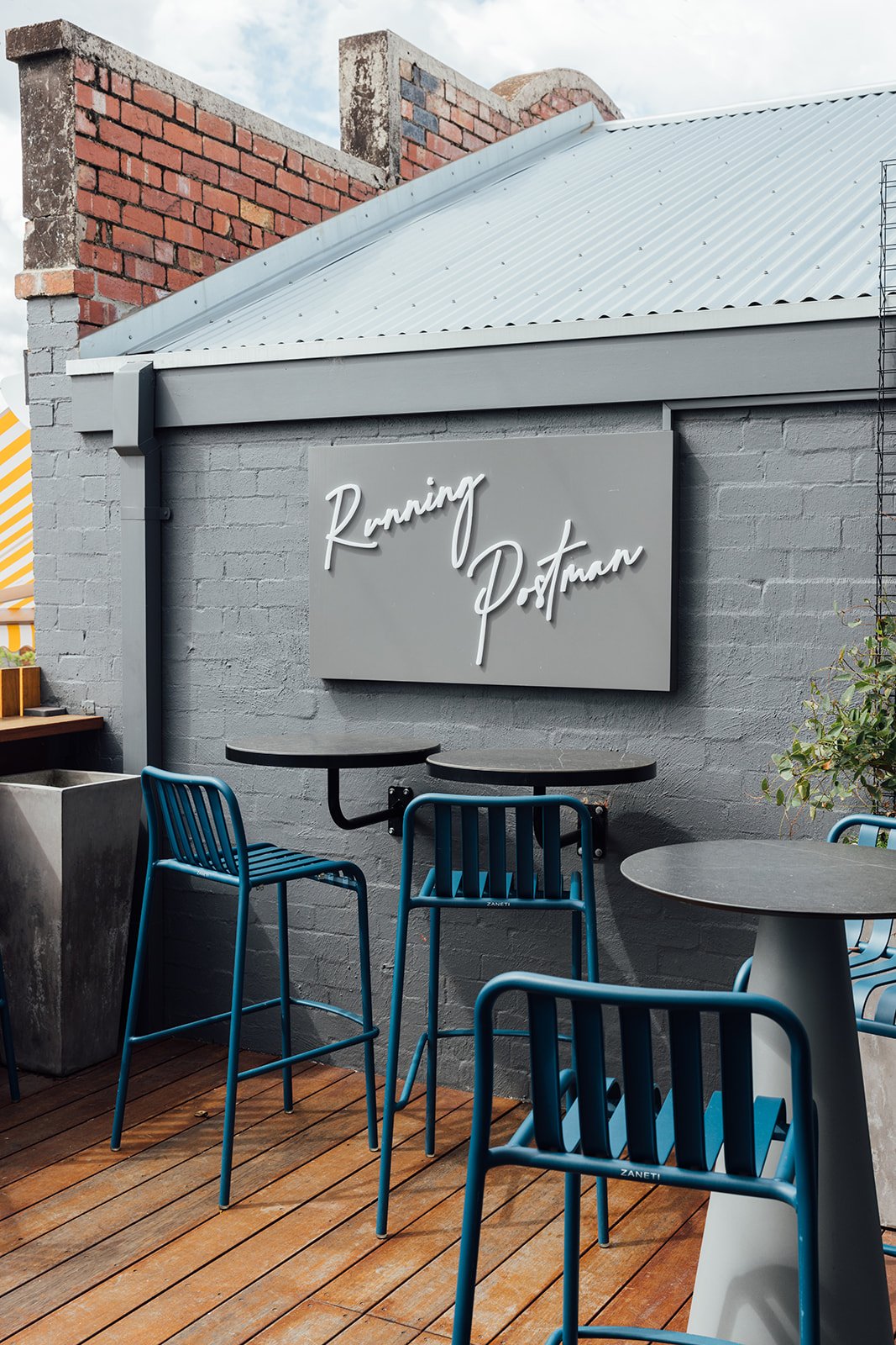 Outdoor area of an event space used by Coral & Co. Dark blue stools sit next to dark stools and table tops. A grey sign, reading the name of the wine bar "Running Postman" in white writing sits on a wall which is also grey. 