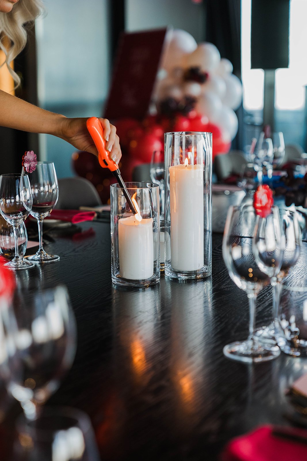 Empty wine glasses with acrylic, scalloped drink tags sit on a dark wooden table around two white candles encased with glass. A lady is lighting the second candle as the other burns. A welcome sign and balloon garland sit blurred in the background behind the table. Styled by Coral & Co.