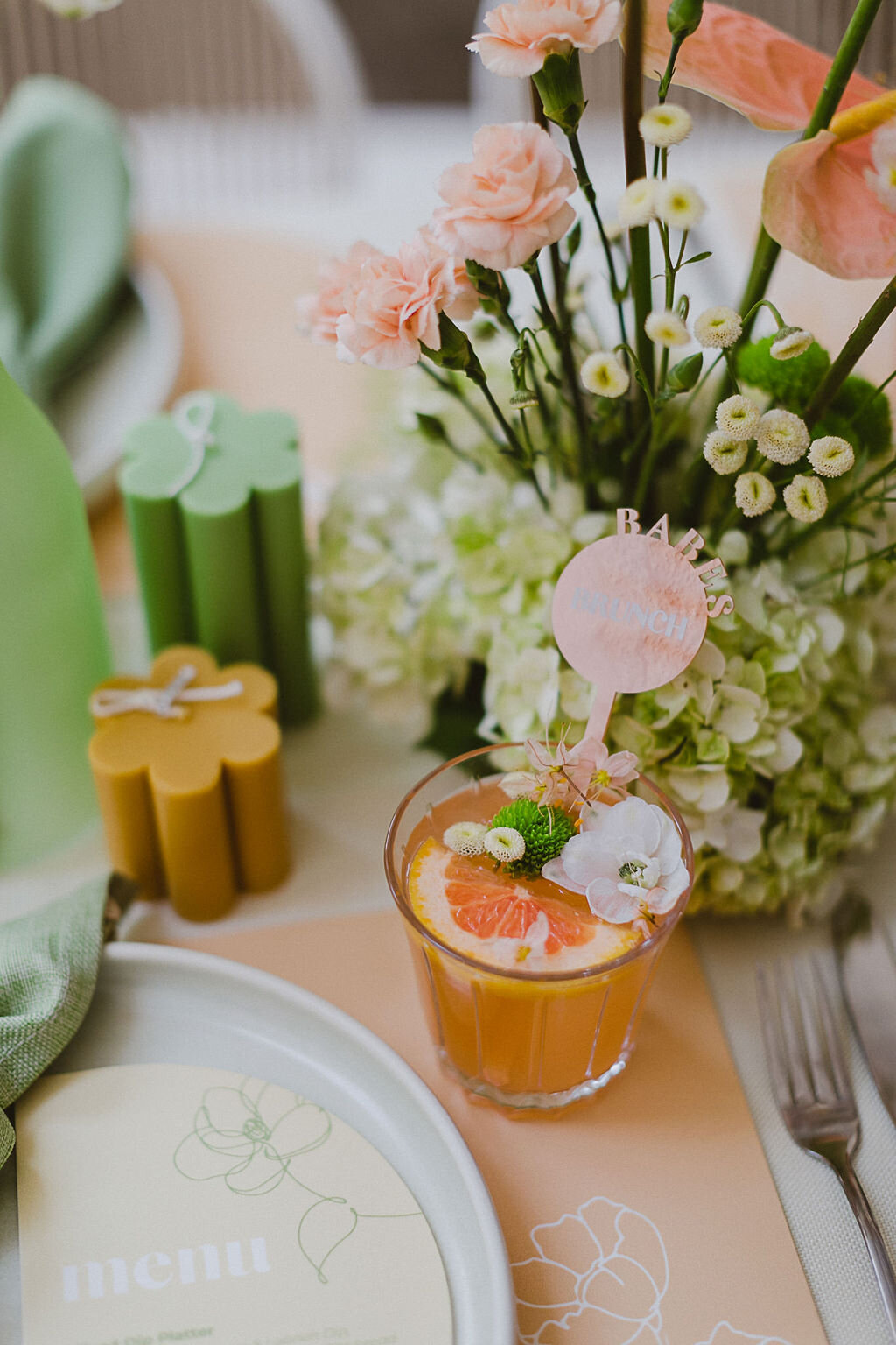 A Melbourne Babes Brunch. A tropical fruit cocktail, with fresh grapefruit and florals in the drinks. A stirrer reads "Babes Brunch" and pink and green florals are in the background next to scalloped unlit candles. 