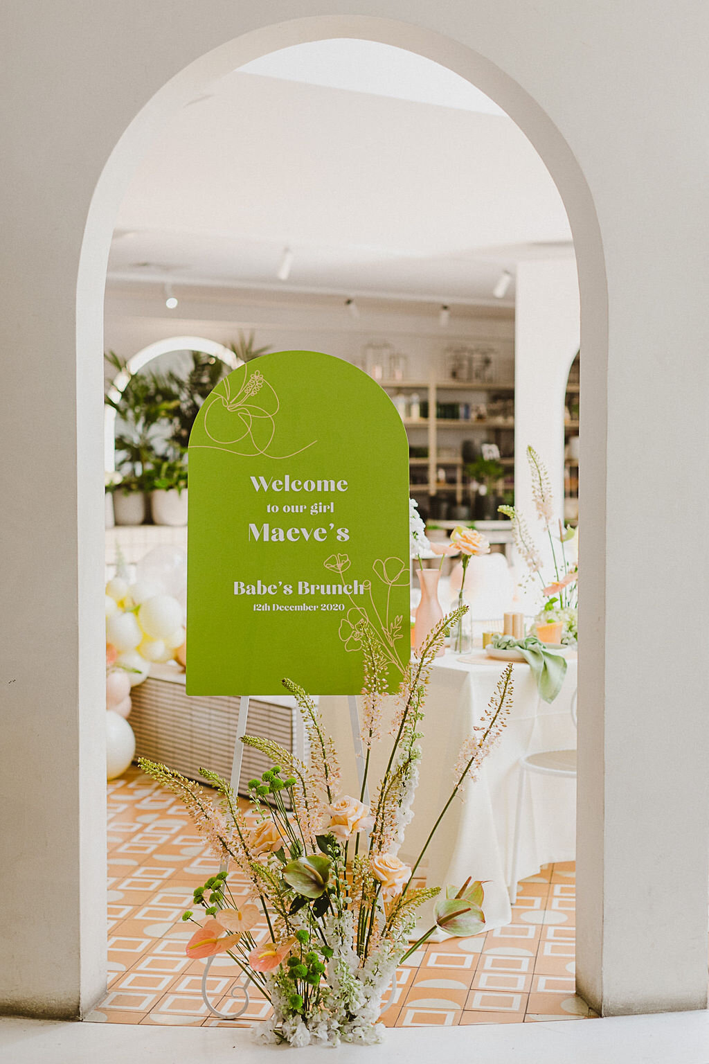 A arch entrance to a dining area, with a green welcome sign and bright, tall florals in front. In the backgorund there are dressed tables and a balloon garland. Welcome to our girl’s Maeve’s super fun and fresh bridal babe’s brunch! With Zoobibi’s art deco and Middle Eastern vibes, the space is filled with light and curved arches to make a grand entrance. 
