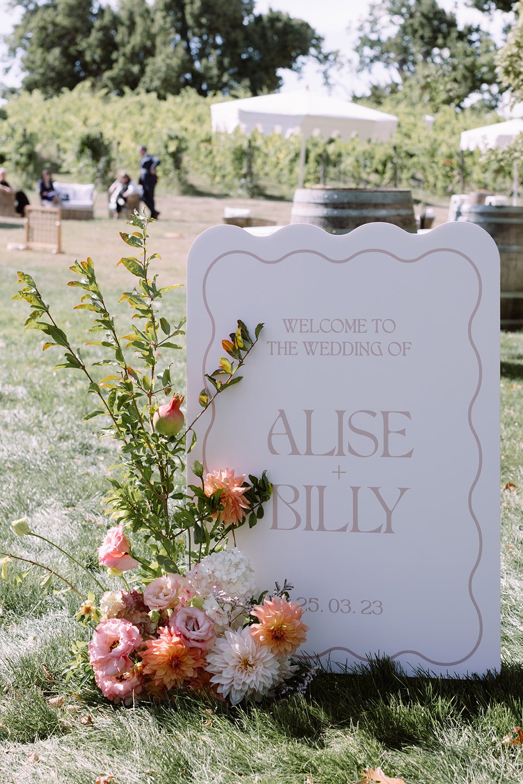 Sitting on grass, a cream, rectangle shape welcome sign with a wavey border, reading "Welcome to the wedding of Alise & Billy", designed by a Melbourne event stylist. A colourful floral setting of pinks, oranges, white and green sits against the left side of the sign. In the background is a game area with old wine barrels, stools and scalloped umbrellas for sitting. 