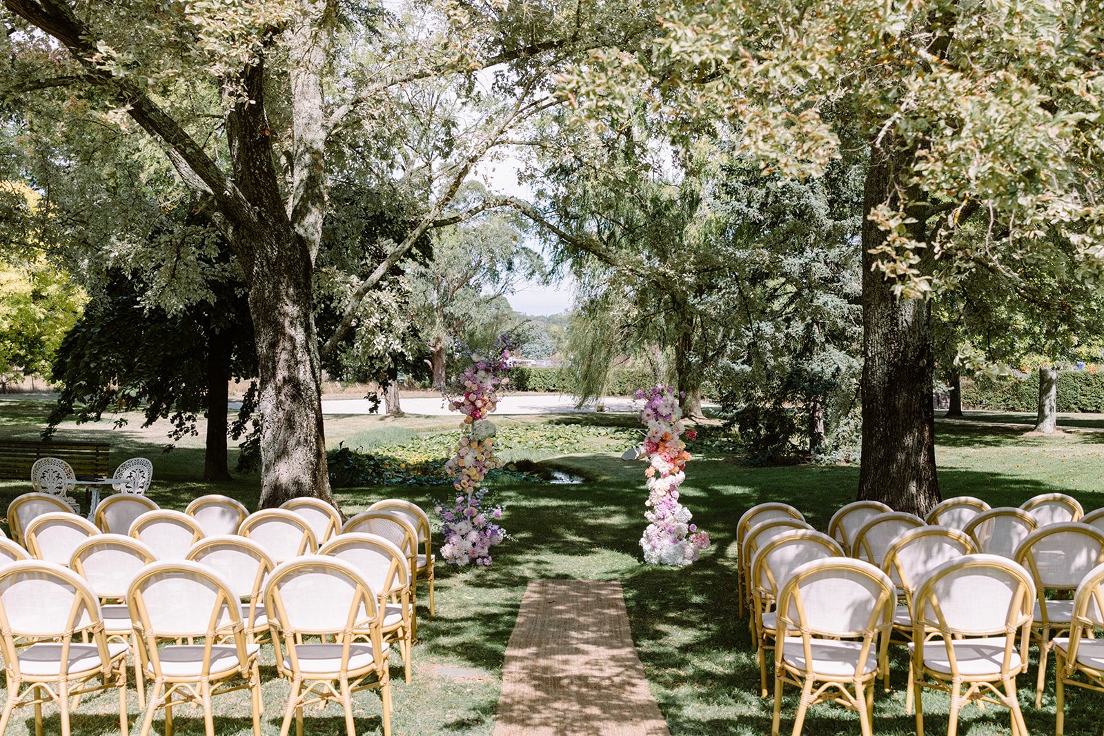 The isle of a wedding at St Agnes Homestead on a bright day. A wedding arch, with bright flowers on both sides sits between two grand trees at the end of the isle. White chairs, sitting on green grass line the isle, facing towards the colourful floral arch. 