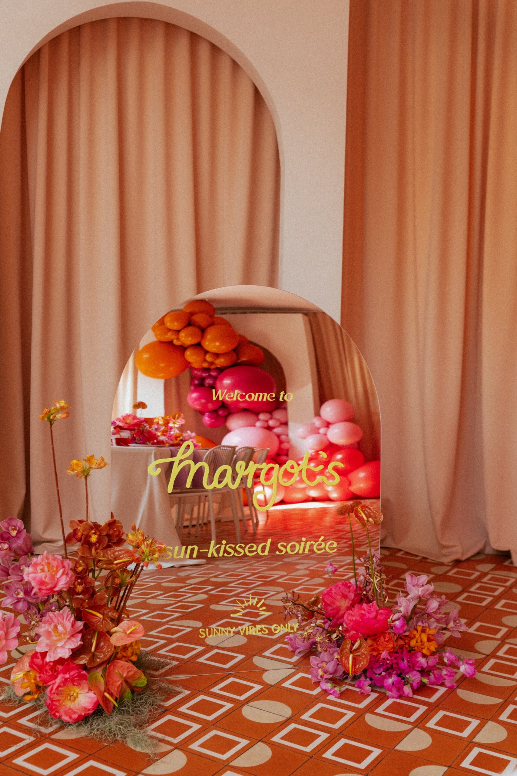 A mirrored arch welcome sign sitting on the floor with two floral arrangements either side. The welcome sign says, "welcome to Margot's sun-kissed-soiree". In the reflection of the sign is fun and popping hot pink and bright orange balloon garland, styled by an event planner in Melbourne. 