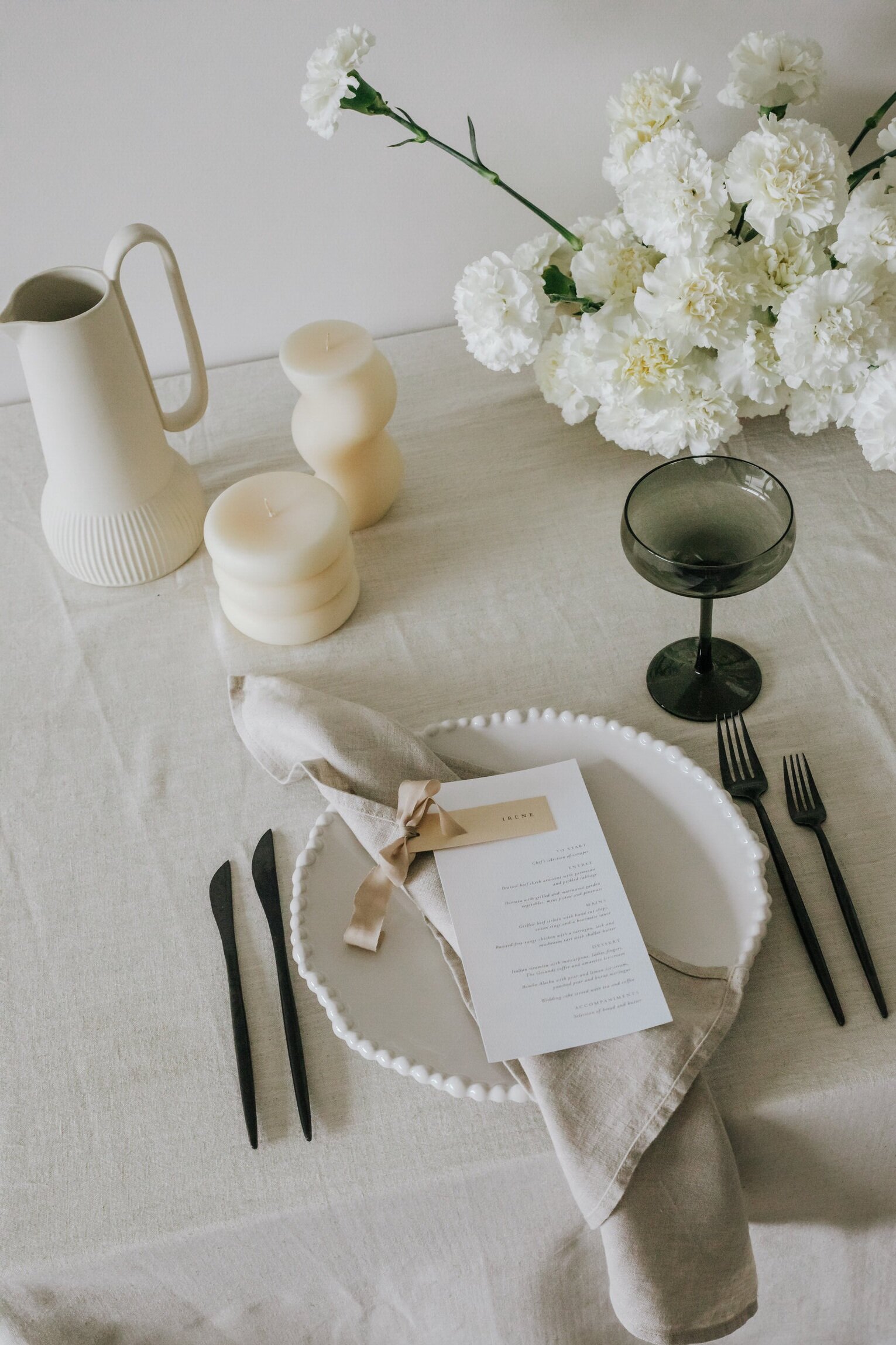A table setting with white florals in the background, next two two candles, both cream and not alight. One wavey and the other shorter and round. A crisp white menu with a cream bow sits on a napkin and plate. An at home dinner setting in Melbourne. 