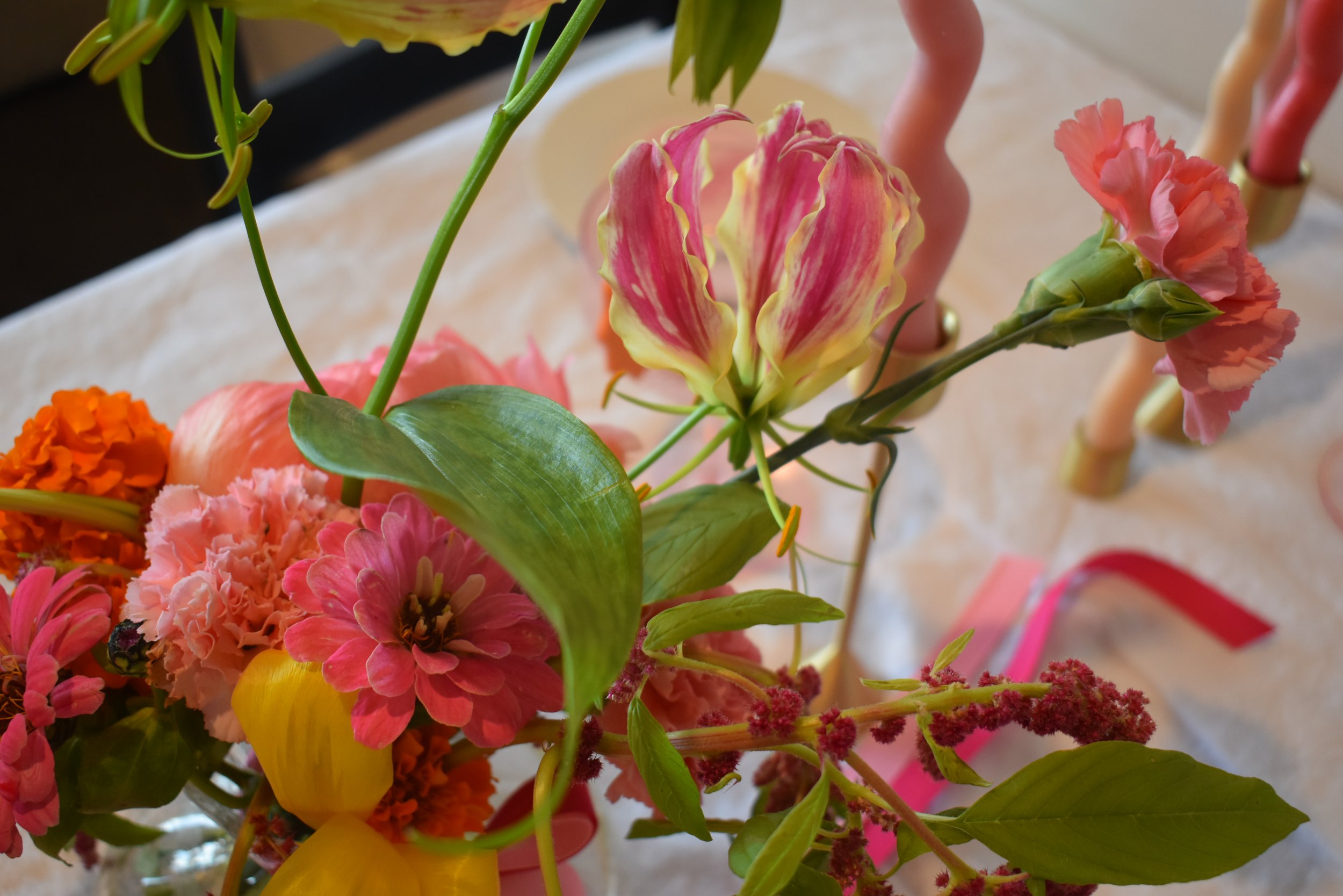 Bright pink, yellow, red and green florals captured from above, looking down on a table setting, with wavey colourful candles in the background on a linen table cloth. 