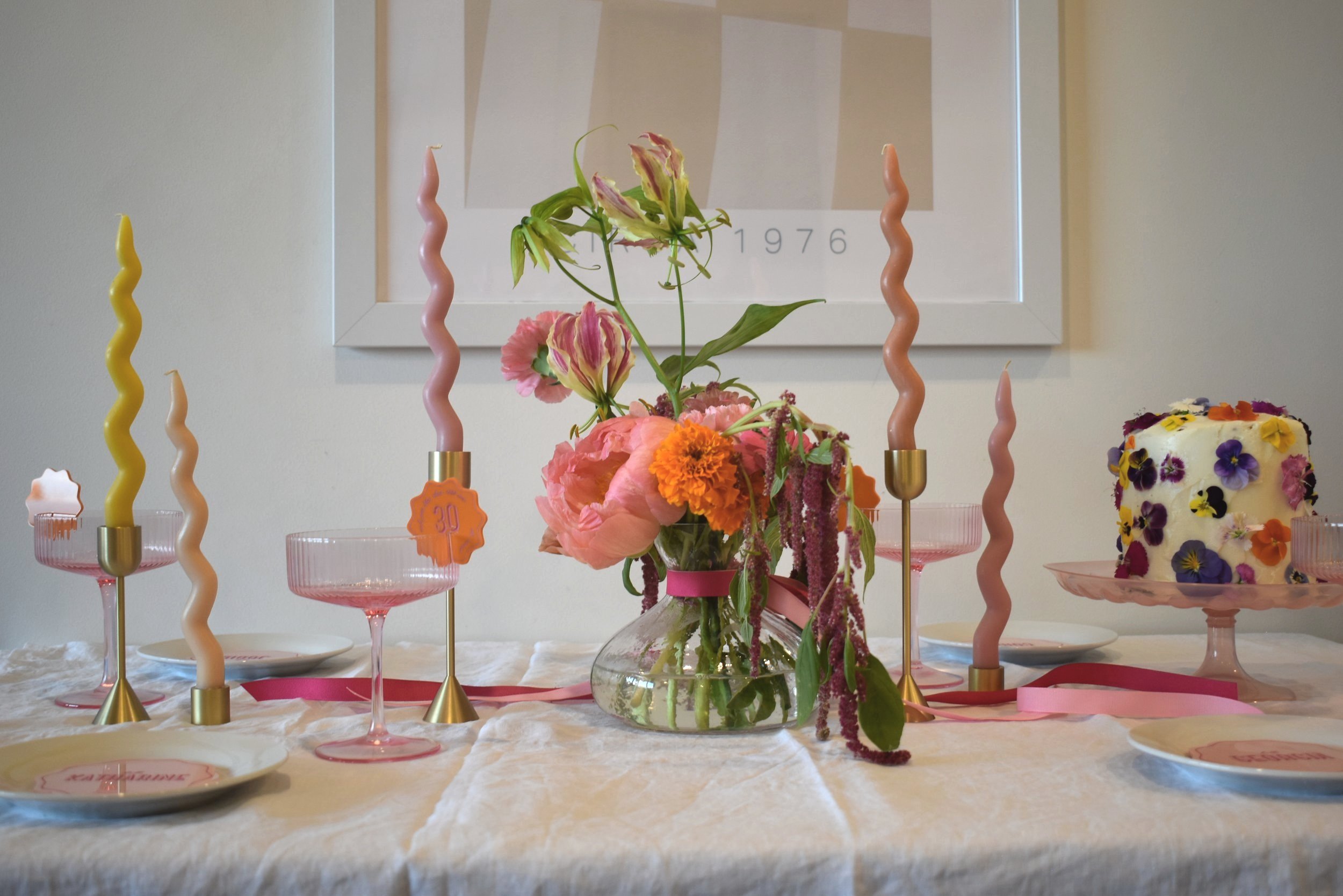 Vibrant table setting, with bright tropical florals in the center, wavey candles with pinks and yellows, sitting next to pink see-through deep martini glasses with orange drinks tags. A cake with florals sits to the right of the table and art sit behind the table on a cream wall. 