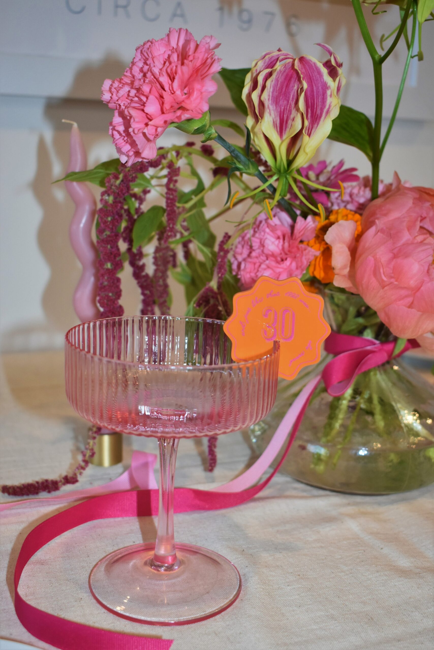 An empty see-through pink martini glass, with a "30" orange drinks tag. Bright pink ribbon flows around the bottom of the glass from the bright & vibrant florals in the background. A pinky purple wavey candle is nestled amongst the florals. A birthday celebration in Melbourne. 
