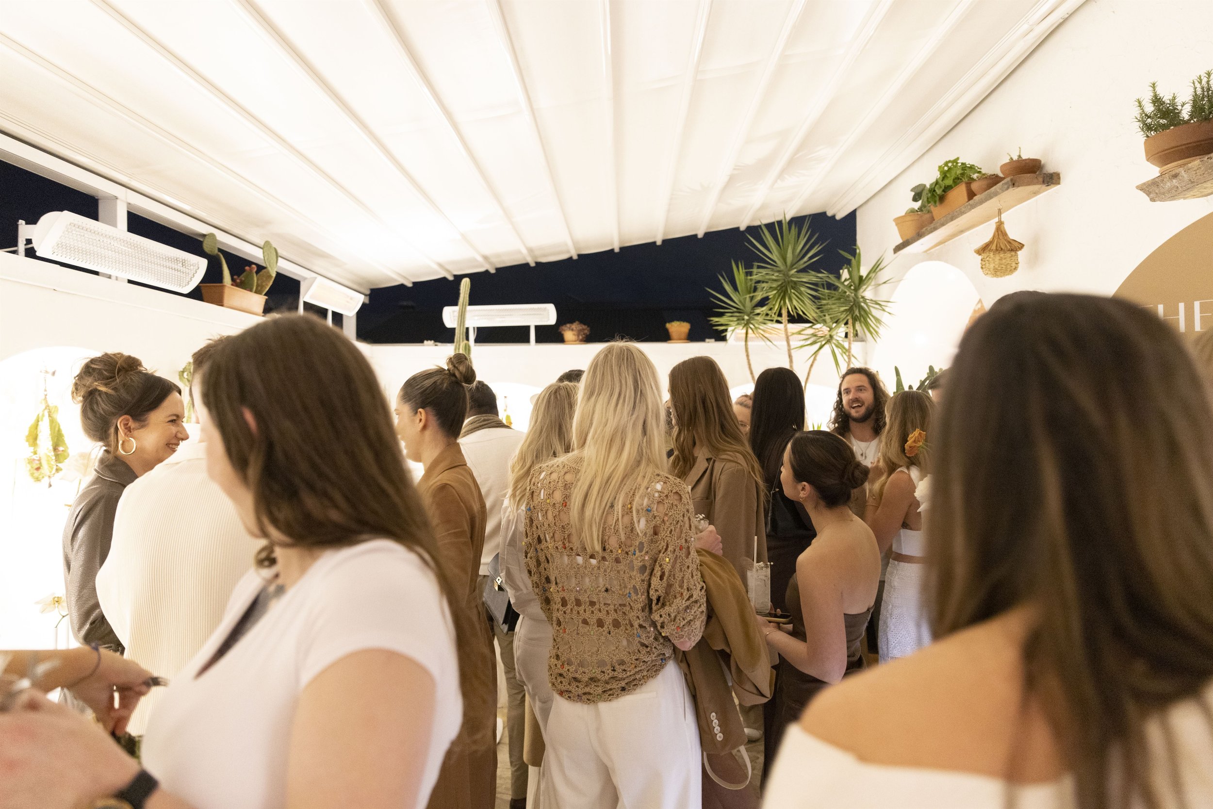 A group of people at an event launch party, styled by Coral and Co. People chat and mingle under a white backdrop, with green florals with the dark night sky peeping through a gap at the end of the room. 