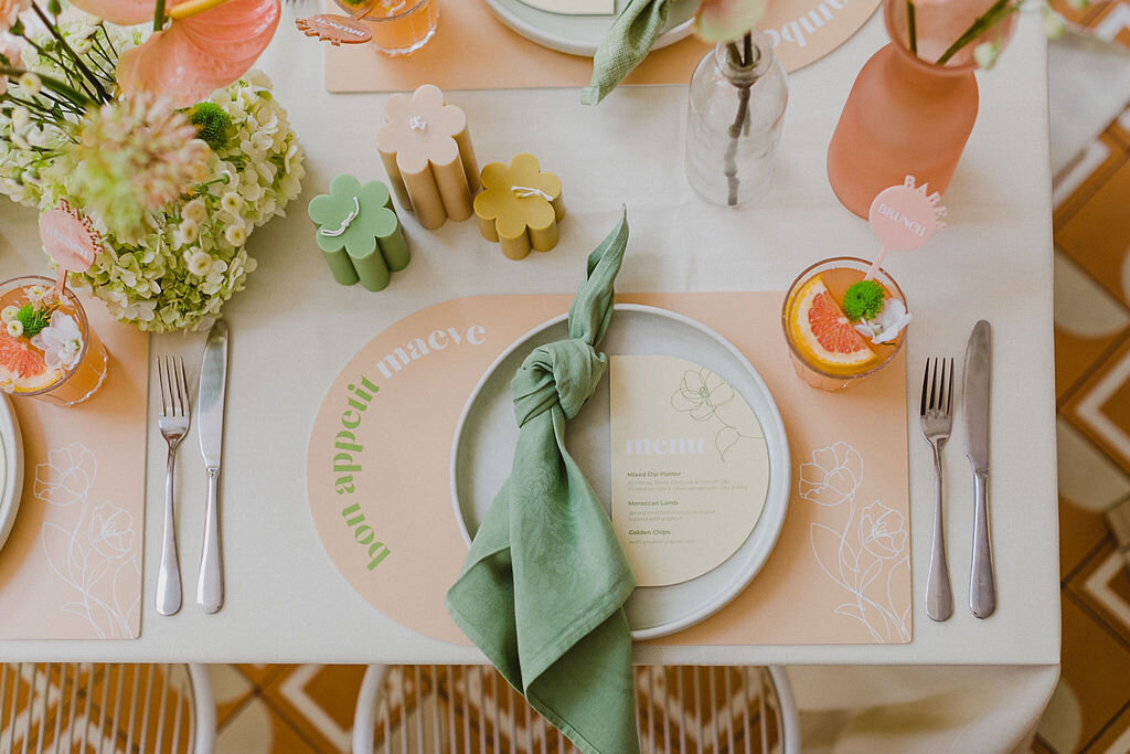 A view from above a table setting. A tropical cocktail with fresh fruit and stirrer sits on top of a curved place setting. A white plate with a curved menu on top sits next to a green tied napkin. Above are scalloped pastel coloured candles, next to florals in the middle of the table. 
We wanted to fill this event with florals from start to finish, so we were delighted when we discovered Aacute and their dazzling daisy pillar candles! Ranging in heights and colours, they added texture and the perfect finish to our table of floral …